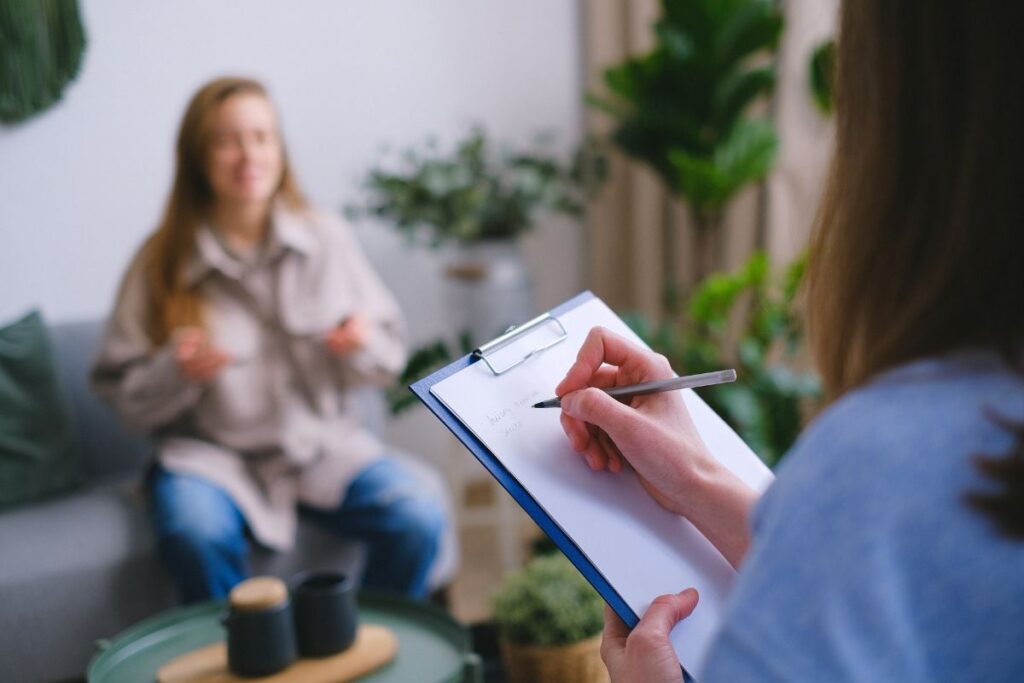 Therapist sitting and taking notes on a blue clipboard with a patient sitting on a gray couch in background