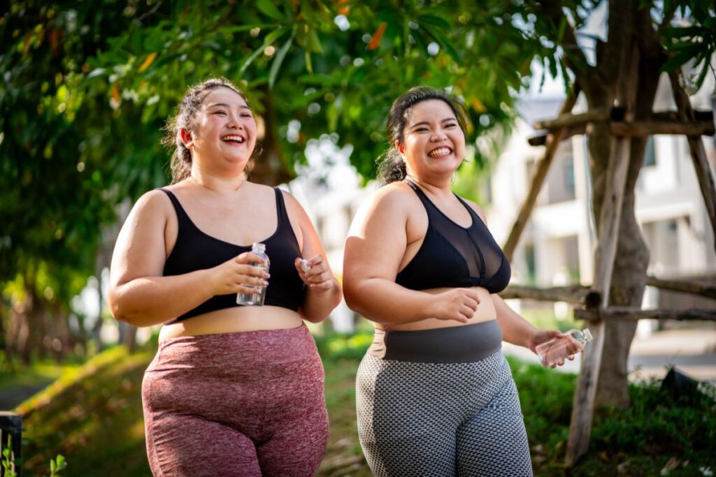 two plus size Asian American women jogging with water bottles in hand and smiling wearing sports bras and leggings with tropical trees in the background