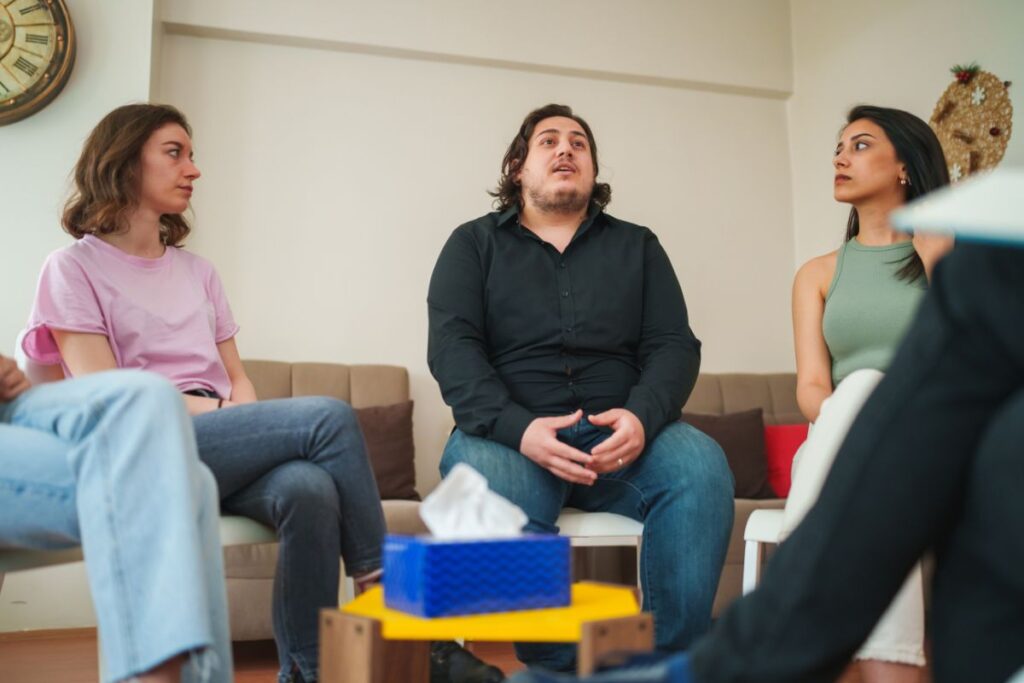 room of people with two females looking to male in the center talking during therapy group 