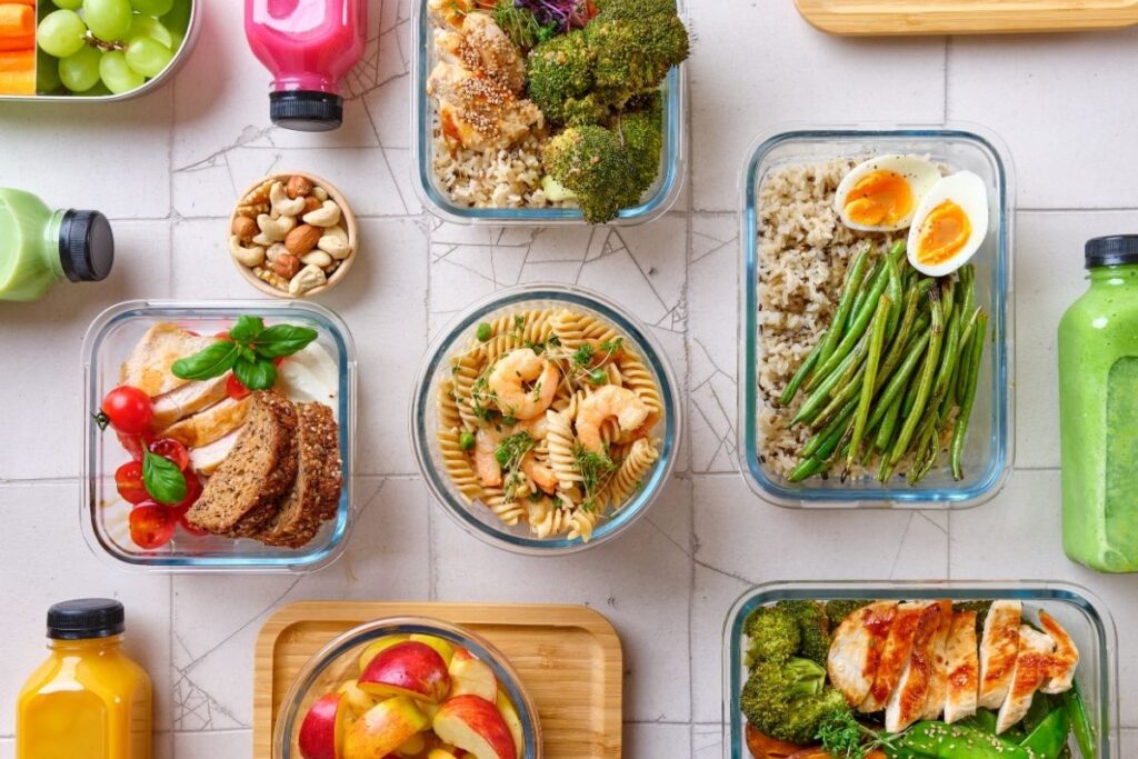 seven glass meal prep containers for balanced meals including eggs, shrimp, rice, green Eans and broccoli with three different smoothies in clear bottles.