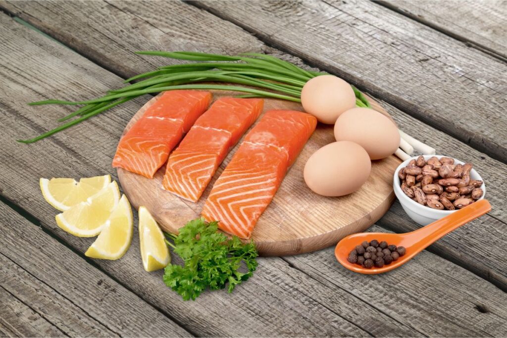 wooden background showing 3 raw salmon filets, a bundle of chives, 4 lemon wedges, a sprig of cilantro, 3 eggs, a small cup of beans and an orange teaspoon of beans 